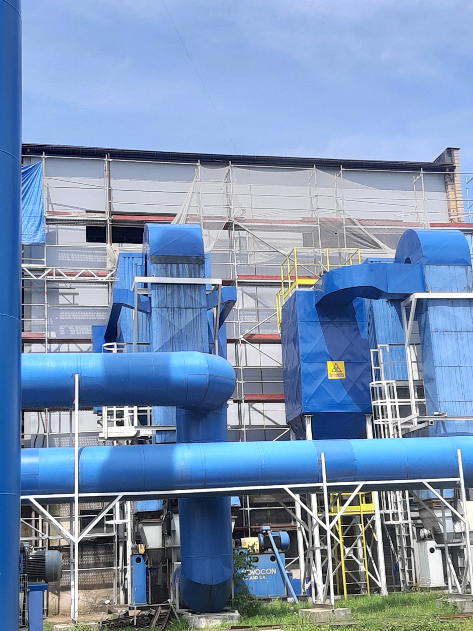 Niebieska instalacja przemysłowa z rurami i urządzeniami, otoczona rusztowaniami i konstrukcją budynku w tle, widoczne ostrzeżenie na jednym z elementów.