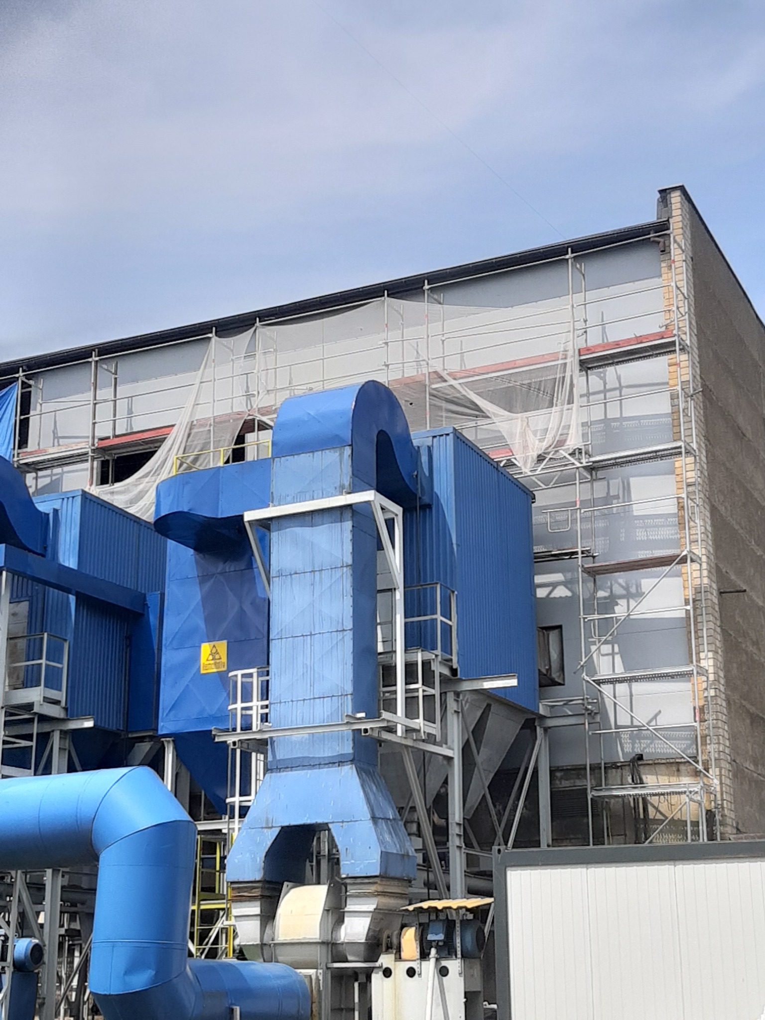 Fragment elewacji budynku przemysłowego z niebieską instalacją wentylacyjną i rusztowaniami, widoczny znak ostrzegawczy.