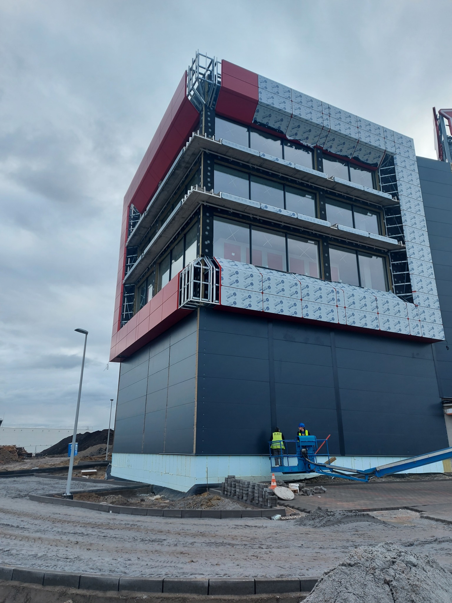 Wielopiętrowy budynek w trakcie montażu elewacji z płyt warstwowych, widoczni dwaj pracownicy na podnośniku koszowym, szaro-bura kolorystyka otoczenia.