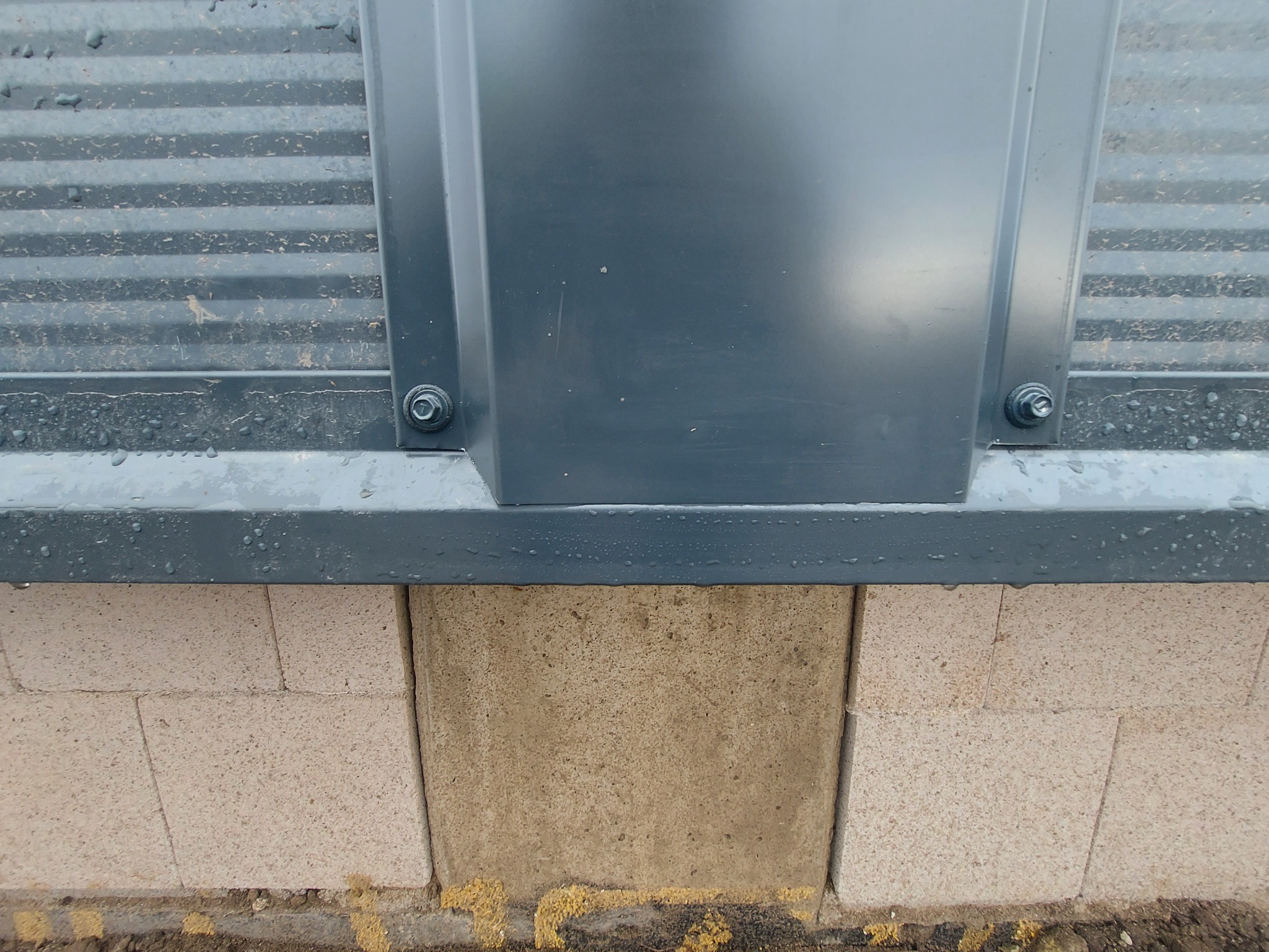 Fragment budynku z szarej blachy falistej i paneli, połączonych śrubami, z widocznymi bloczkami fundamentowymi z betonu komórkowego.