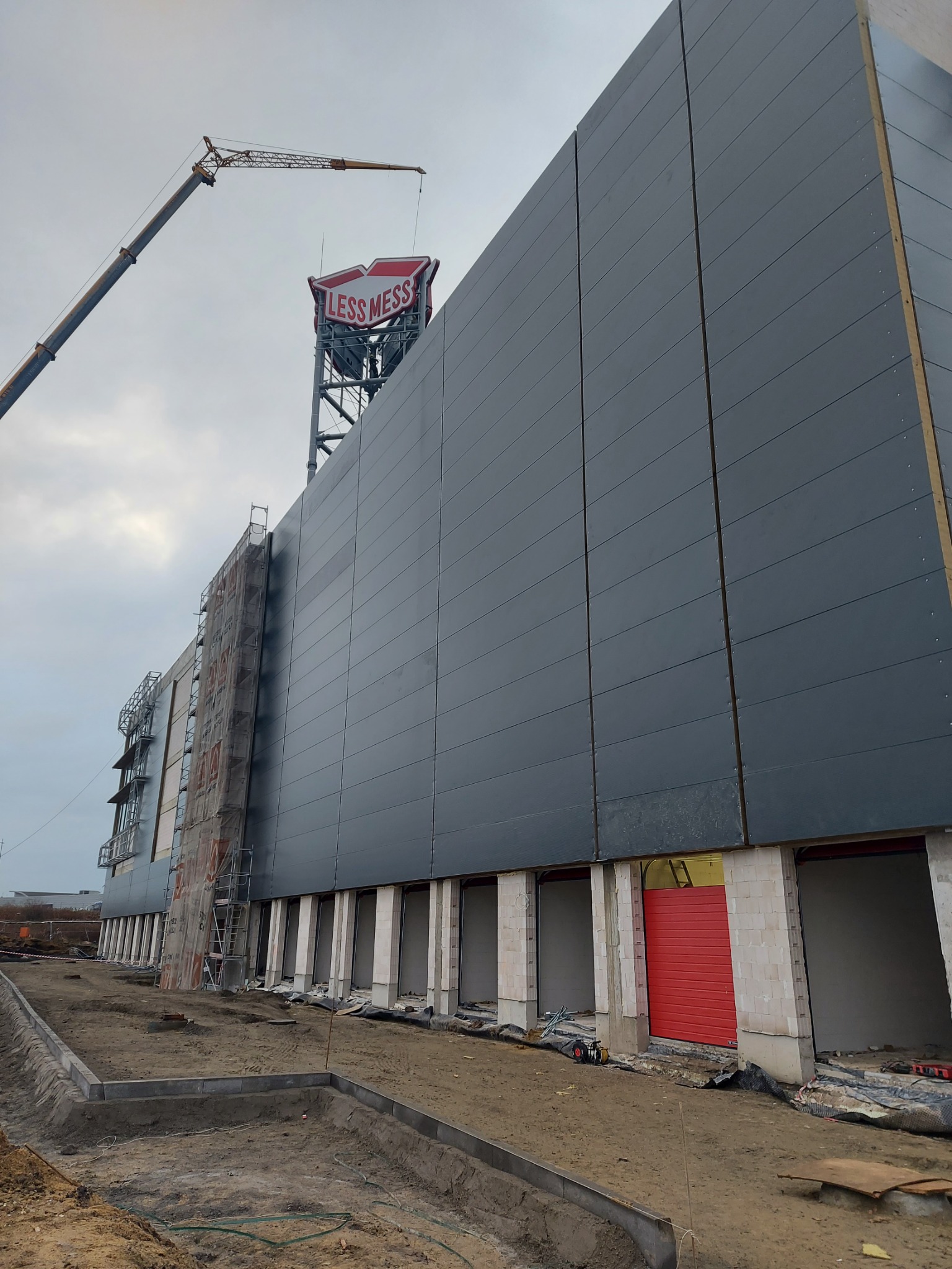 Wznoszona hala przemysłowa z szarymi panelami warstwowymi, widoczny dźwig budowlany i logo 'LESS MESS' na szczycie konstrukcji.