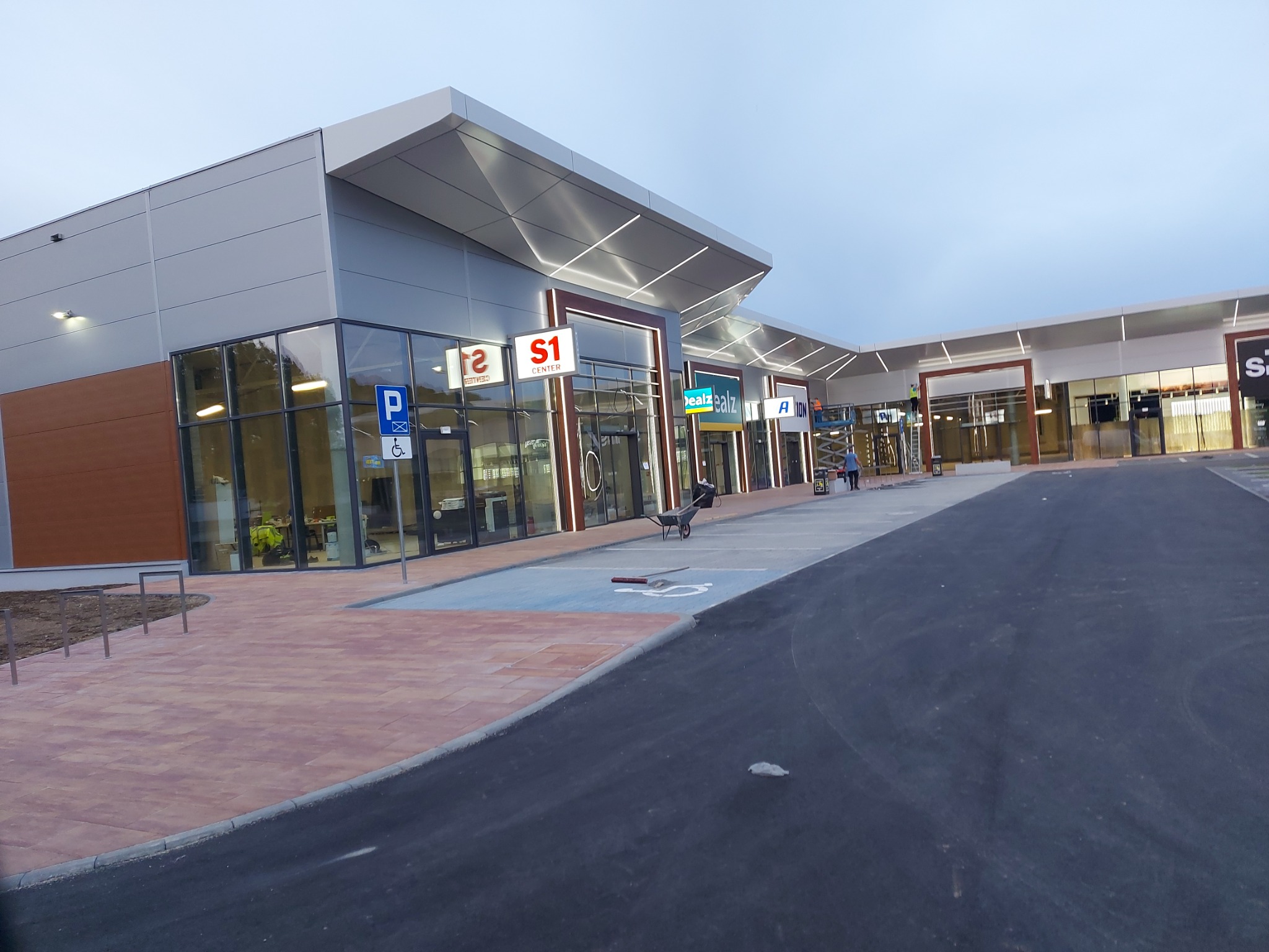 Nowoczesny budynek hali handlowej z elementami szarości i drewna, parkingiem z miejscami dla niepełnosprawnych, widoczne prace wykończeniowe i osoby w oddali.