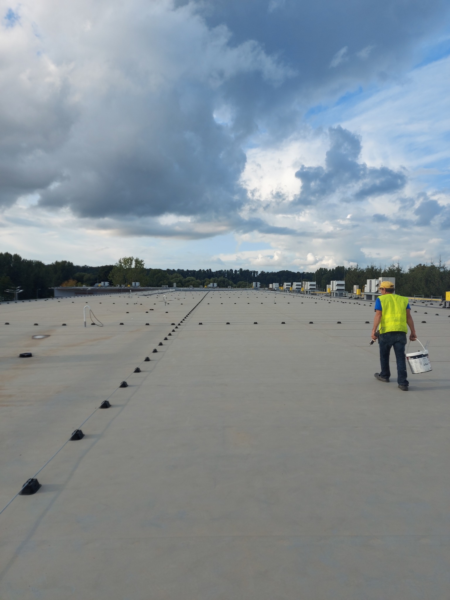 Pracownik w żółtej kamizelce i czapce z wiadrem farby na rozległym, płaskim dachu hali w Międzyrzecu Podlaskim, widoczne liczne punkty mocowań i systemy wentylacyjne w tle pod pochmurnym niebem.