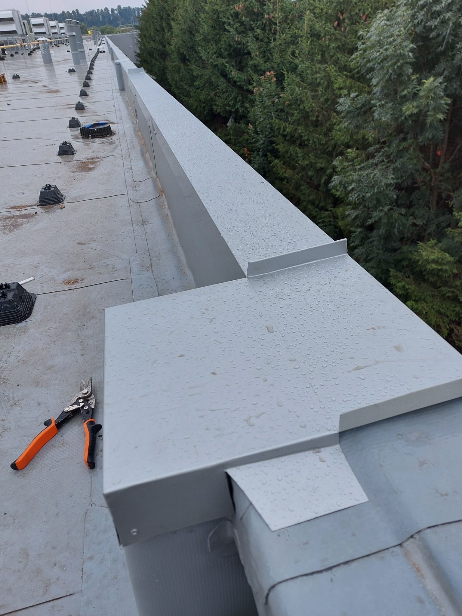 Montaż obróbek blacharskich na dachu hali, widoczne nożyce do blachy z pomarańczowymi uchwytami i krople deszczu na powierzchni blachy.