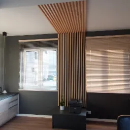 Wnętrze biura z oknami przesłoniętymi poziomymi osłonami okiennymi w odcieniach beżu, drewniane lamele dekoracyjne na ścianie i suficie, ciemnoszare ściany i meble w jasnych odcieniach szarości.