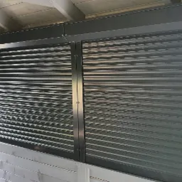 Zewnętrzne rolety metalowe w kolorze grafitowym, zamontowane w altanie ogrodowej, częściowo zasłonięte, widoczny fragment drewnianej ławki i trawnik w tle.