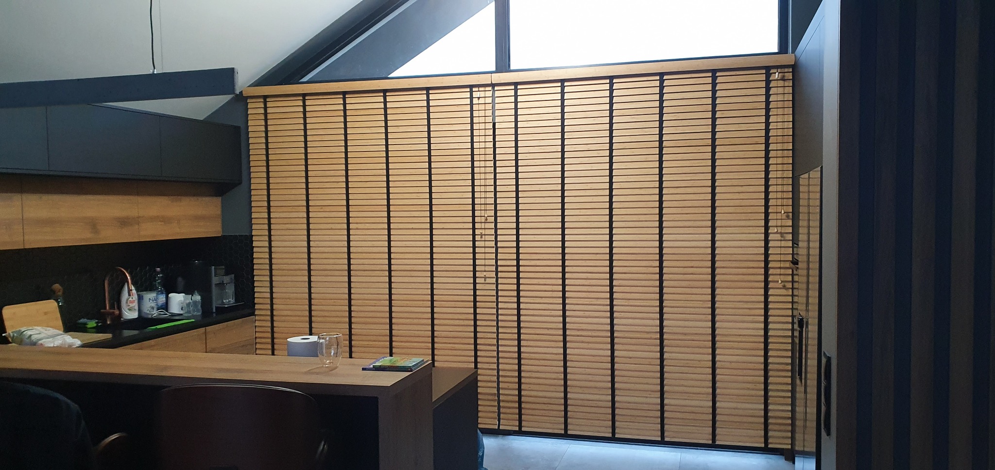 Jasne, drewniane żaluzje poziome zamontowane w oknie o nietypowym kształcie, widoczne w nowoczesnej kuchni z drewnianymi blatami i ciemnymi szafkami.