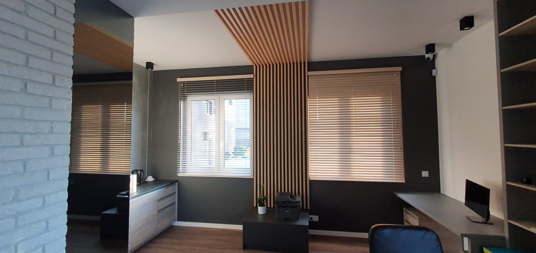 Wnętrze biura z oknami przesłoniętymi poziomymi osłonami okiennymi w odcieniach beżu, drewniane lamele dekoracyjne na ścianie i suficie, ciemnoszare ściany i meble w jasnych odcieniach szarości.