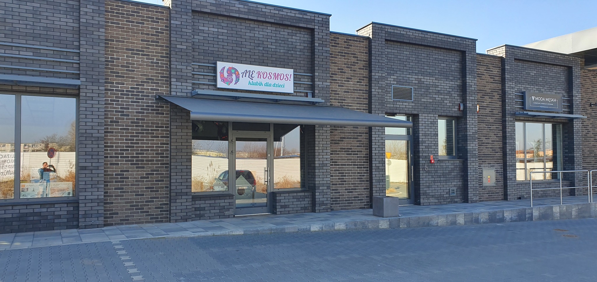 Elewacja budynku z szarej cegły z zamontowaną szarą markizą nad wejściem do lokalu 'Ale Kosmos! Klubik dla dzieci'.