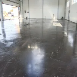 Świeżo wylana, gładka posadzka przemysłowa w hali, z widocznymi refleksami światła i niewielkimi smugami wody na powierzchni, otwartymi drzwiami w tle i widocznym sprzętem budowlanym na zewnątrz.