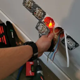 Elektryk sprawdza napięcie w gniazdku za pomocą próbnika napięcia, widoczne przewody, puszka instalacyjna, zamontowane gniazdka, poziomica Milwaukee, wkrętarka Metabo i torba narzędziowa Wera