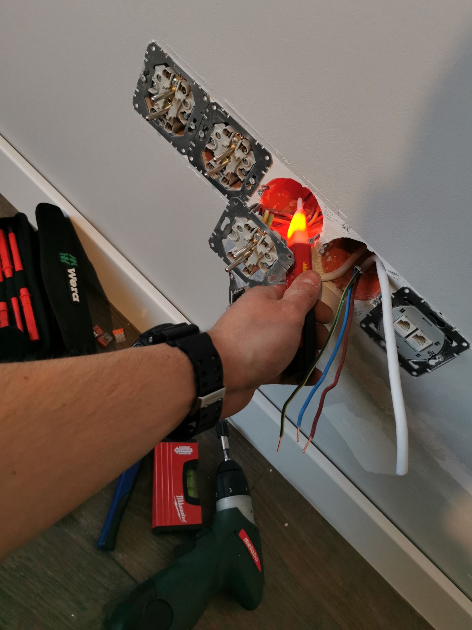 Elektryk sprawdza napięcie w gniazdku za pomocą próbnika napięcia, widoczne przewody, puszka instalacyjna, zamontowane gniazdka, poziomica Milwaukee, wkrętarka Metabo i torba narzędziowa Wera