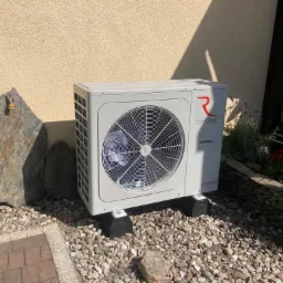 Szara jednostka zewnętrzna z logo Rotenso, umieszczona na betonowych wspornikach na żwirowym podłożu, obok dużego kamienia i rabaty kwiatowej.