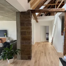 Jasne panele podłogowe w przestronnym wnętrzu z drewnianymi belkami stropowymi i ceglanym filarem, widok na salon z kanapą i telewizorem.