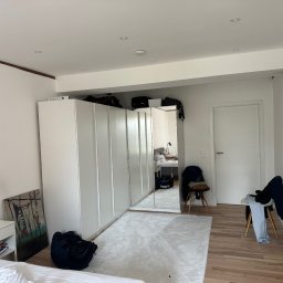 Usługi Budowlano - Wykończeniowe Mikołaj Kida - Biała szafa wnękowa z lustrzanymi drzwiami w jasnym pokoju. Widoczne drewniane panele podłogowe i biały dywan. Na szafie torby, na podłodze torba podróżna.