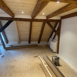 Usługi Budowlano - Wykończeniowe Mikołaj Kida - Poddasze z drewnianą konstrukcją stropu i częściowo wykończonymi ścianami. Widoczne elementy przygotowane do dalszych prac wykończeniowych, takie jak listwy i farby.