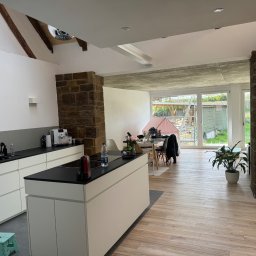 Usługi Budowlano - Wykończeniowe Mikołaj Kida - Nowoczesna kuchnia z wyspą, drewnianą podłogą i kamiennymi elementami. Widoczne białe szafki, czarny blat i duże okno z widokiem na zewnątrz. Oświetlenie punktowe.