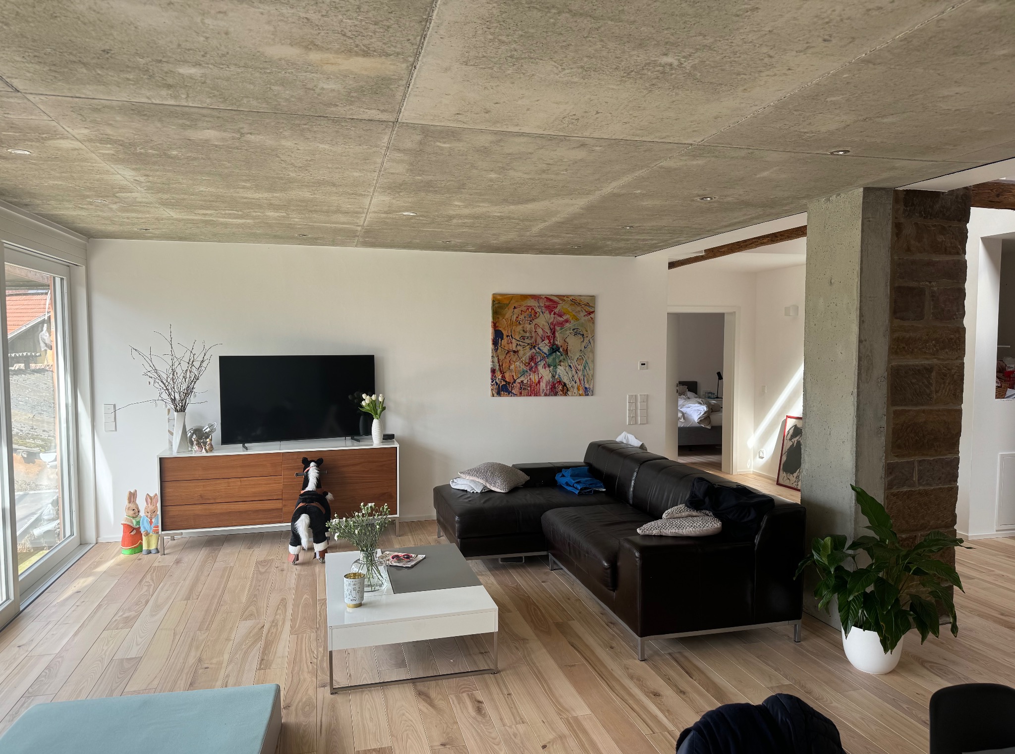 Nowoczesny salon z surowym, betonowym sufitem, jasnymi panelami podłogowymi i ciemną skórzaną sofą. Widoczny element dekoracyjny z motywem wielkanocnym.