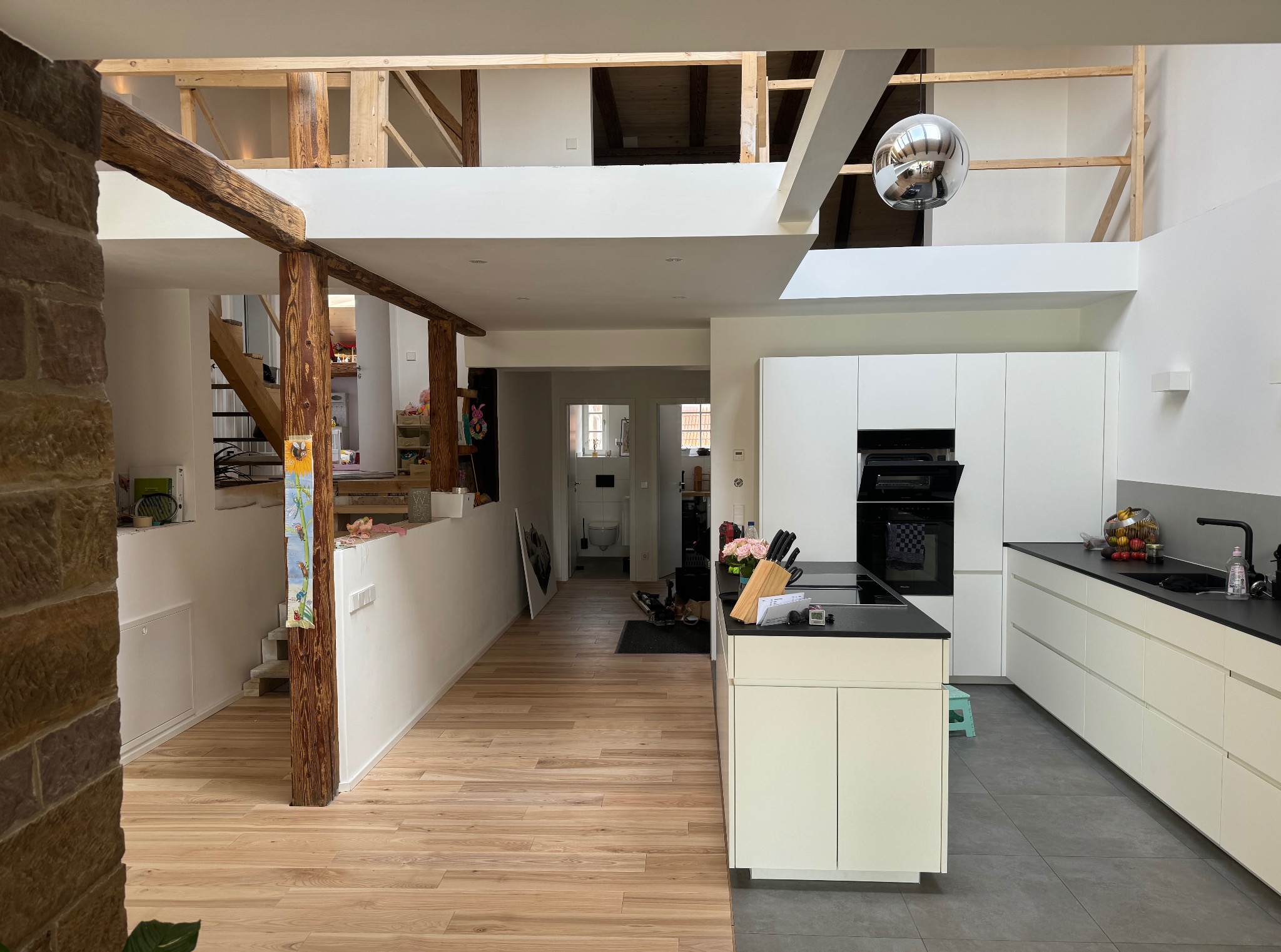 Przestronne wnętrze domu po remoncie: jasna kuchnia z czarnym blatem, drewniana podłoga, schody z drewnianymi belkami. Widoczna otwarta przestrzeń i wysoki sufit.