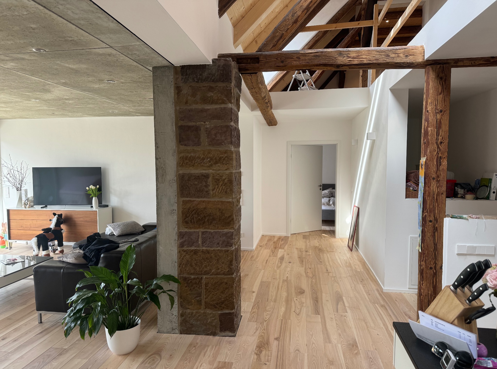 Jasne panele podłogowe w przestronnym wnętrzu z drewnianymi belkami stropowymi i ceglanym filarem, widok na salon z kanapą i telewizorem.