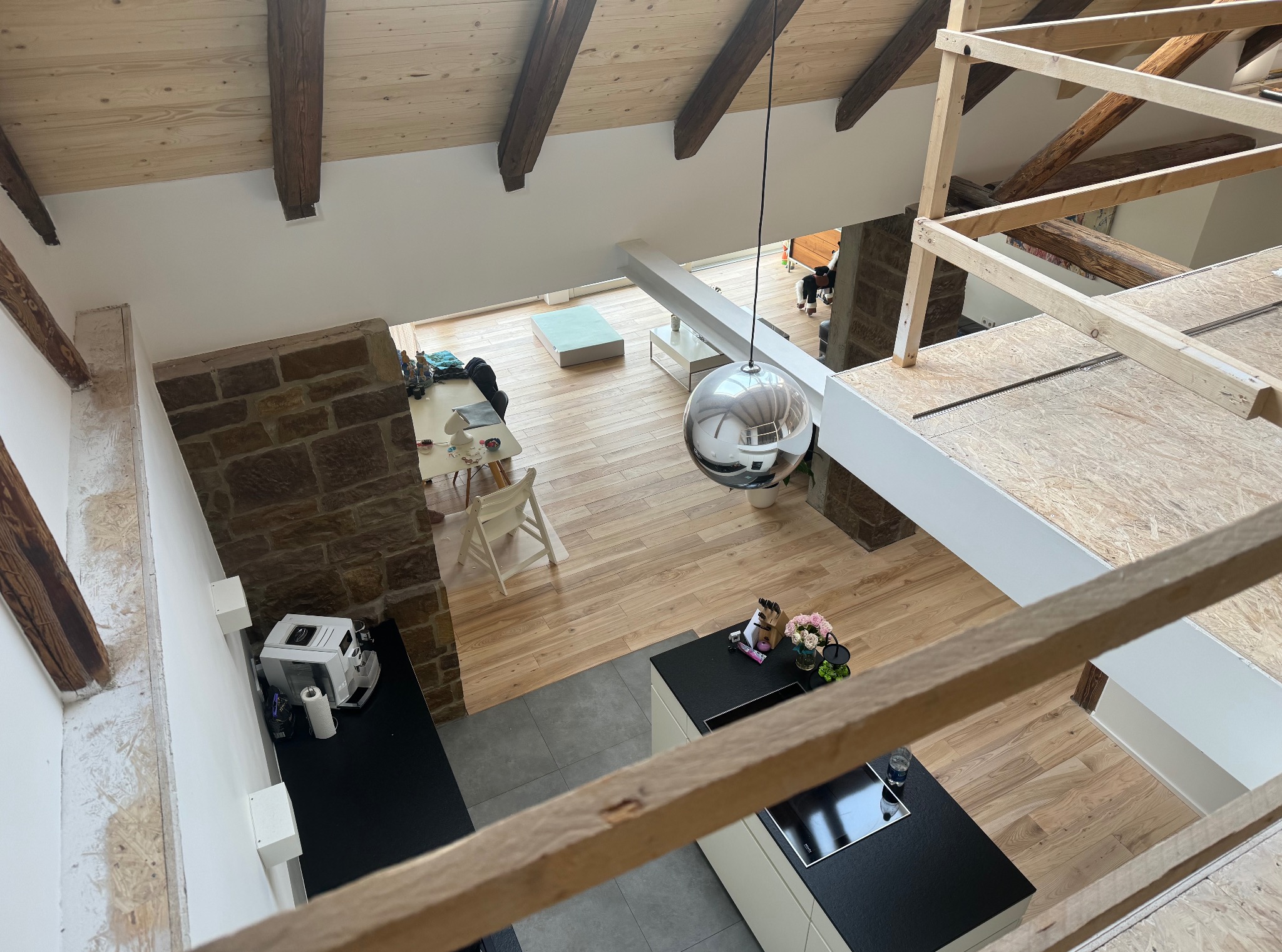 Wnętrze domu w trakcie wykańczania z drewnianymi belkami stropowymi, widok z góry na kuchnię, salon i konstrukcję antresoli. Widoczne elementy wykończeniowe.