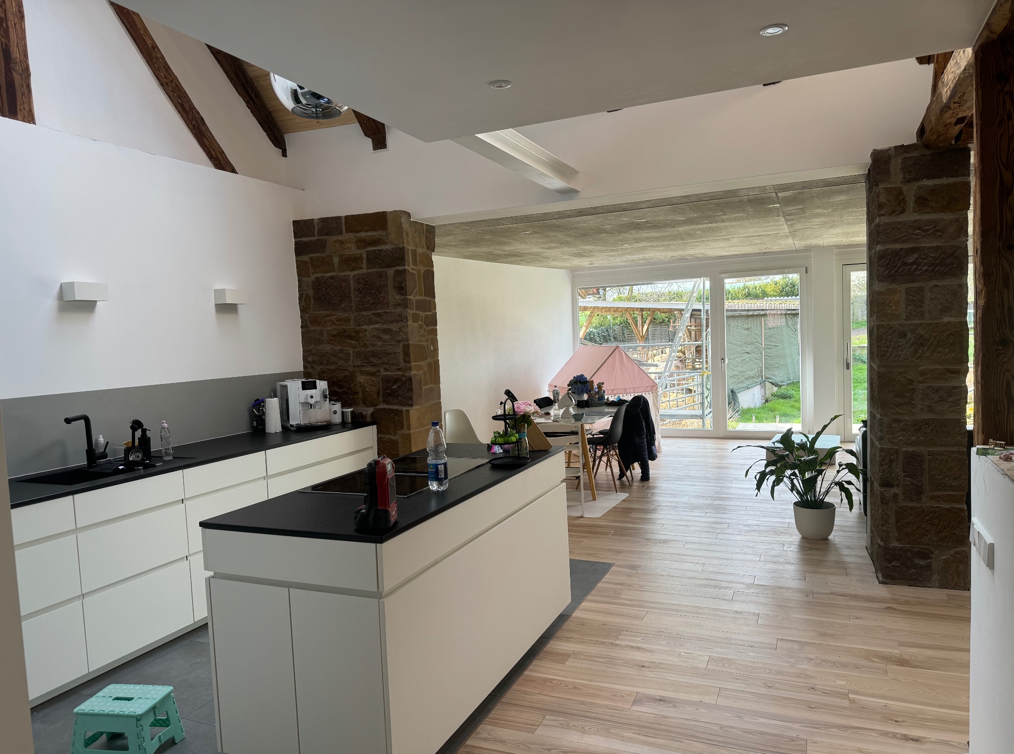 Nowoczesna kuchnia z wyspą, drewnianą podłogą i kamiennymi elementami. Widoczne białe szafki, czarny blat i duże okno z widokiem na zewnątrz. Oświetlenie punktowe.