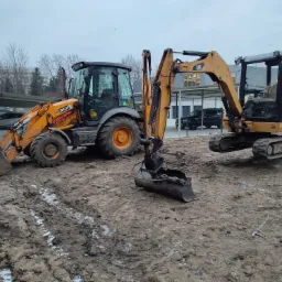 Dwa żółte pojazdy budowlane: koparko-ładowarka JCB 3CX i minikoparka CAT 302.5C, stojące na ubłoconym placu budowy z widocznymi śladami opon i resztkami śniegu.