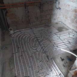 CREATIVE WORLD ART SPÓŁKA Z OGRANICZONĄ ODPOWIEDZIALNOŚCIĄ - Montaż ogrzewania podłogowego w surowym pomieszczeniu z ceglanymi ścianami, widoczne rury grzewcze na folii z siatką, mocowane czarnymi uchwytami.