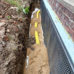 CREATIVE WORLD ART SPÓŁKA Z OGRANICZONĄ ODPOWIEDZIALNOŚCIĄ - Wykop wzdłuż fundamentu budynku z widoczną folią kubełkową, izolacją termiczną i żółtymi rurami w piasku.