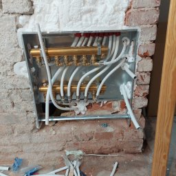 CREATIVE WORLD ART SPÓŁKA Z OGRANICZONĄ ODPOWIEDZIALNOŚCIĄ - Rozdzielacz ogrzewania podłogowego w metalowej skrzynce, zamontowany w surowej ścianie z cegły, częściowo wypełnionej pianką montażową, widoczne rury i elementy instalacyjne, na podłodze resztki...