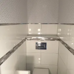 Nowoczesna, mała toaleta z białymi płytkami i dekoracyjnym pasem z mozaiki, wisząca muszla klozetowa z chromowanym przyciskiem, umywalka narożna i tapeta w subtelny wzór.