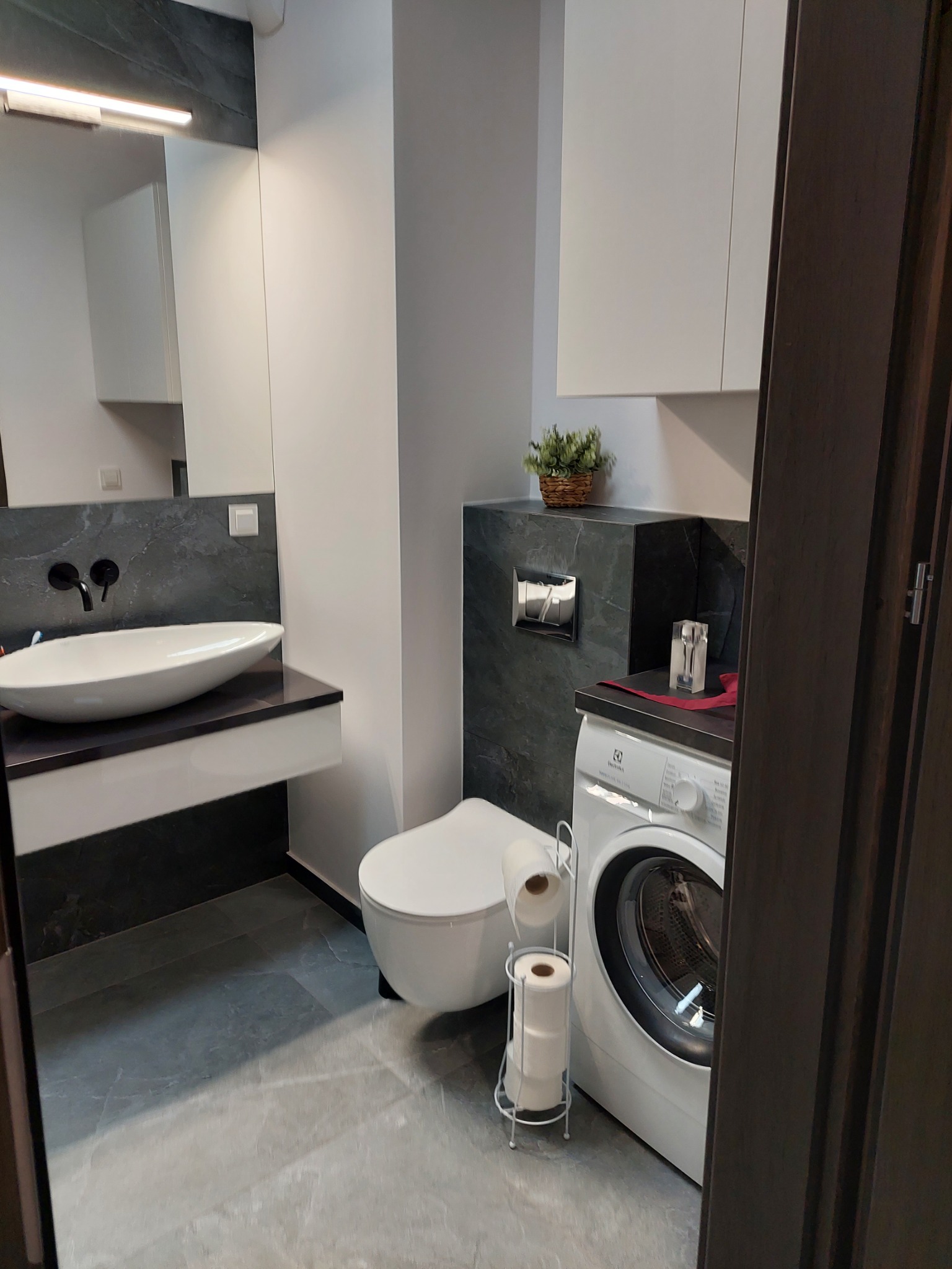 Nowoczesna łazienka z umywalką nablatową na czarnym blacie, wiszącą toaletą i pralką Electrolux z akcesoriami, wykończona szarą płytką imitującą marmur.