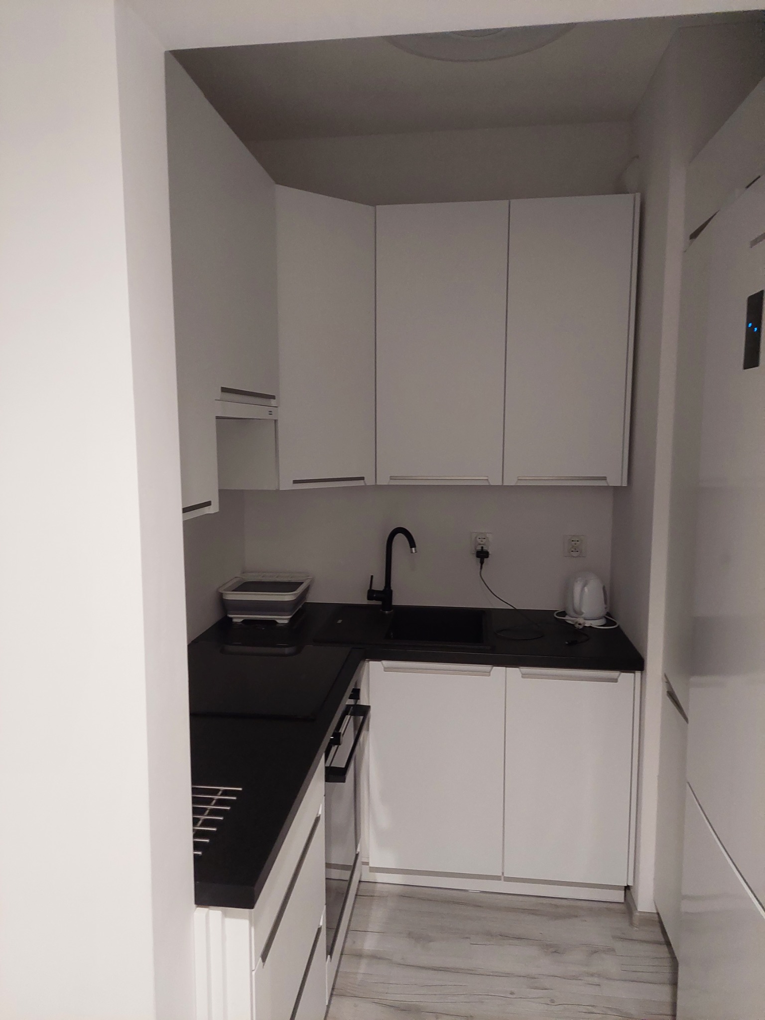 Nowoczesna, minimalistyczna kuchnia z białymi szafkami i czarnym blatem, widok na zlew z czarną armaturą, biały czajnik elektryczny i szarą podłogę.