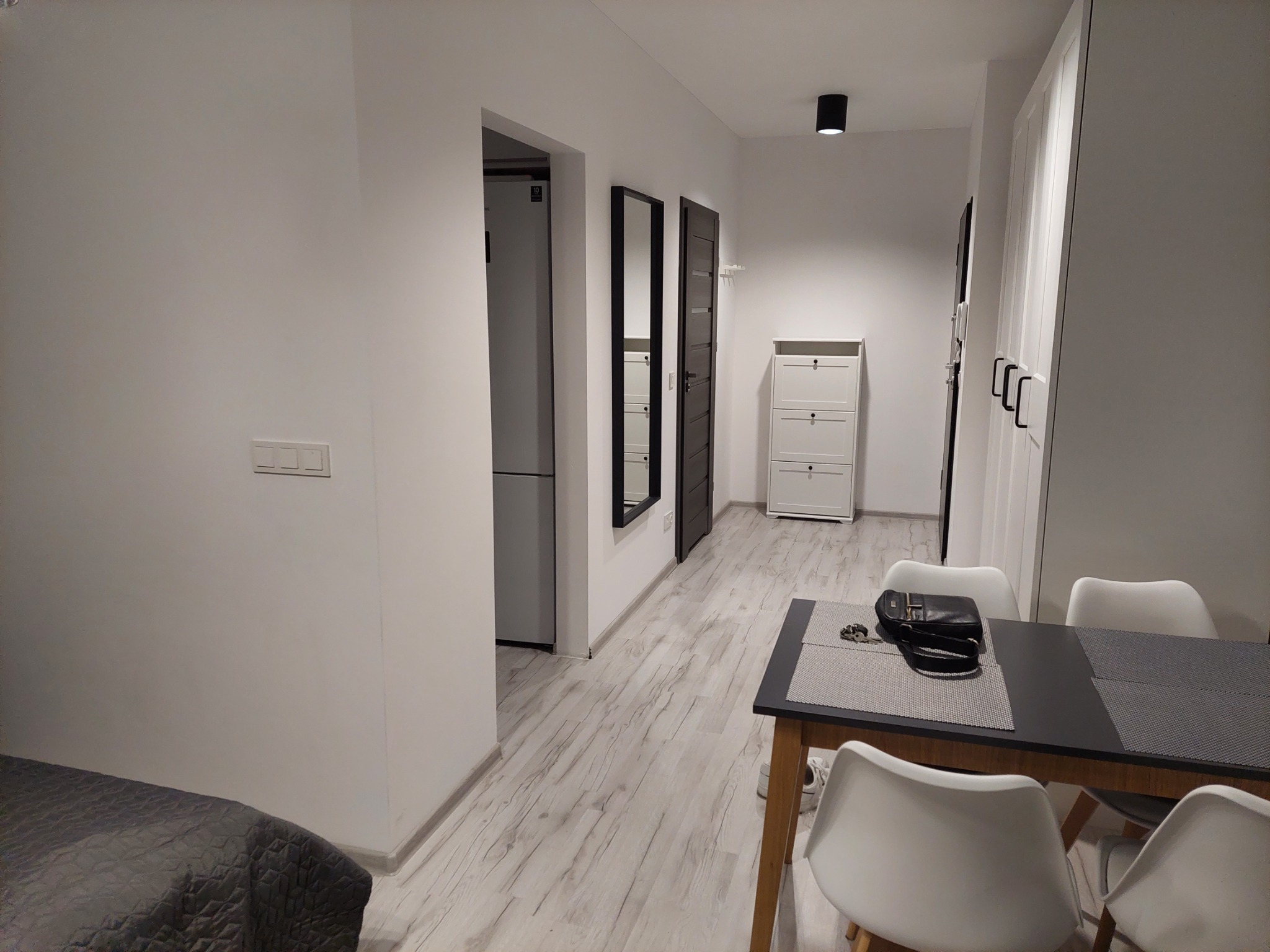 Jasne wnętrze mieszkania z podłogą imitującą drewno, czarnym stołem jadalnym z akcesoriami i białymi krzesłami, widok na przedpokój z lustrem i szafką na buty.