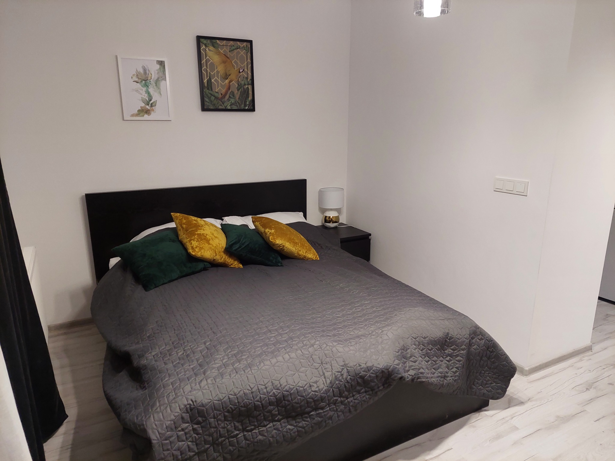 Sypialnia z łóżkiem z czarnym zagłówkiem, szarą pikowaną narzutą i dekoracyjnymi poduszkami w kolorach zieleni i złota. Na ścianie wiszą dwa obrazy w ramkach.