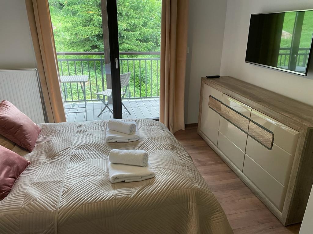 Jasna sypialnia z wyjściem na balkon z widokiem na las. Na łóżku leżą złożone białe ręczniki, a obok stoi komoda z telewizorem na ścianie.