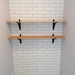 Dwie drewniane półki zamontowane na białej ścianie z cegły w Rychtalu, wsparte na czarnych, metalowych wspornikach.