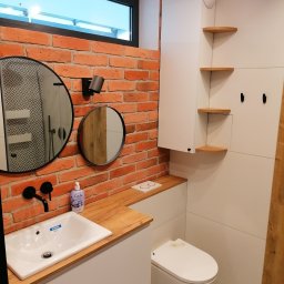 Adrian Świerczyński - Nowoczesna łazienka z umywalką nablatową na drewnianym blacie, ceglaną ścianą, okrągłymi lustrami i wiszącą toaletą na tle wzorzystej podłogi.