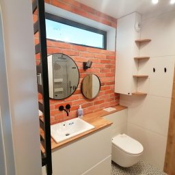 Adrian Świerczyński - Nowoczesna łazienka z ceglaną ścianą, okrągłymi lustrami, drewnianym blatem i białą armaturą, podkreślona geometrycznym wzorem na podłodze.