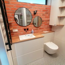 Adrian Świerczyński - Nowoczesna łazienka z ceglaną ścianą, okrągłymi lustrami i drewnianym blatem pod umywalką, wzorzysta podłoga.