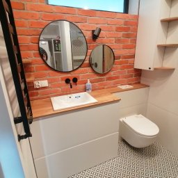 Adrian Świerczyński - Nowoczesna łazienka z ceglaną ścianą, okrągłymi lustrami, białą umywalką nablatową na drewnianym blacie, podwieszaną toaletą i wzorzystą podłogą.