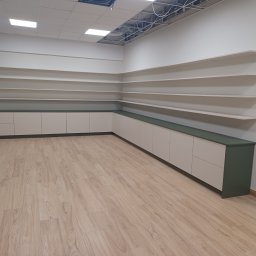 Adrian Świerczyński - Długa szafka z beżowymi frontami i ciemnozielonym blatem, ciągnąca się wzdłuż ściany, z rzędem półek nad nią, na jasnej podłodze imitującej drewno, w pomieszczeniu z częściowo odsłoniętym sufitem.