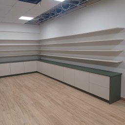 Adrian Świerczyński - Półki ścienne i szafki z zielonym blatem w jasnym pomieszczeniu z drewnianą podłogą, widoczne niedokończone elementy sufitu podwieszanego.