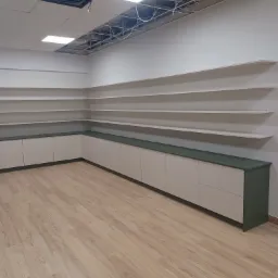 Półki ścienne i szafki z zielonym blatem w jasnym pomieszczeniu z drewnianą podłogą, widoczne niedokończone elementy sufitu podwieszanego.