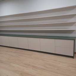 Adrian Świerczyński - Długi rząd szafek z jasnymi frontami i zielonym blatem, zamontowany wzdłuż ściany z półkami, na podłodze panele imitujące drewno. Widoczne elementy sufitu podwieszanego.