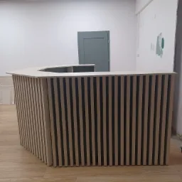 Beżowa lamelowa lada recepcyjna o heksagonalnym kształcie, ustawiona na drewnianej podłodze, widok z przodu na tle jasnych ścian w trakcie wykańczania.