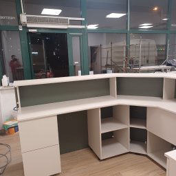 Adrian Świerczyński - Recepcja na wymiar w trakcie montażu, widoczne puste półki i szuflady, tło stanowią okna witrynowe z widocznym rusztowaniem i odbiciem osoby.