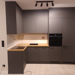 Adrian Świerczyński - Nowoczesna, szara zabudowa kuchenna z drewnianym blatem, czarnym piekarnikiem i czarną płytą grzewczą, oświetlona reflektorami punktowymi.