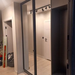 Adrian Świerczyński - Wnętrze przedpokoju z szafą wnękową z przesuwnymi drzwiami lustrzanymi w ciemnej ramie. Widoczny poziomica oparta o ścianę i worek ze śmieciami.