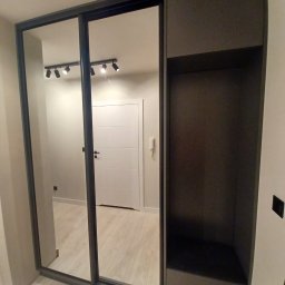 Adrian Świerczyński - Dwudrzwiowa szafa wnękowa z lustrami, czarna rama, obok otwarta półka z ciemnoszarą tapetą wewnątrz, widok na białe drzwi w tle.